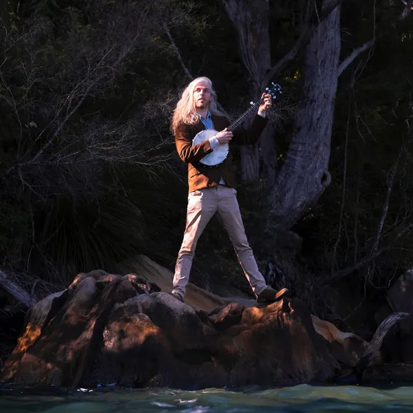 Home album cover: the banjo player standing on sandstone at the water's edge, bush behind